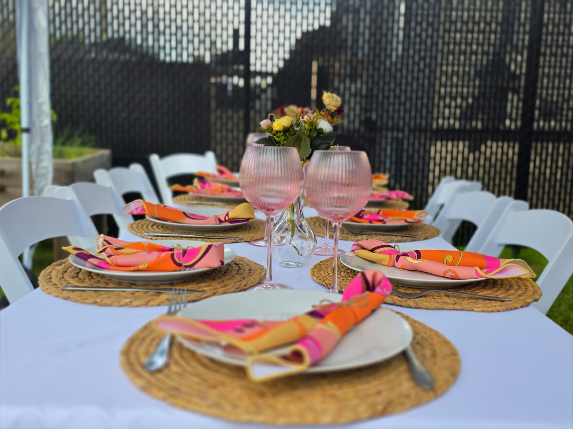 Table dressée en plein air avec chaises Martha blanches, napperons en osier, serviettes colorées et verres roses